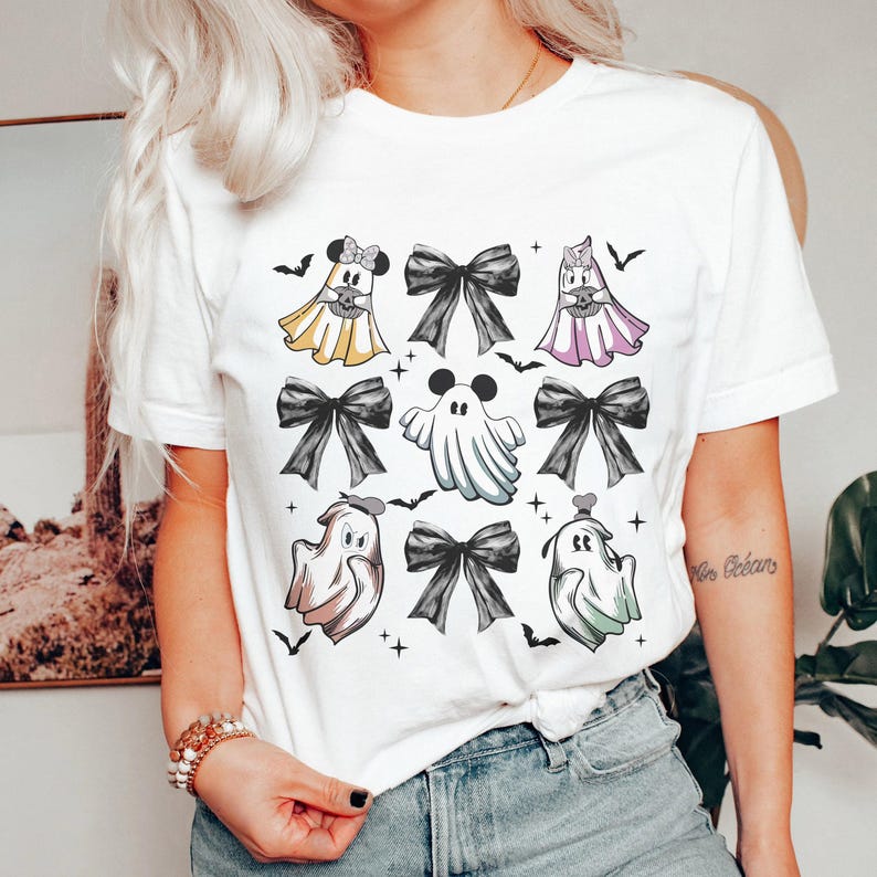 May include: White t-shirt featuring a Halloween-themed design. The design includes cartoon ghosts, black bows, bats, and stars. The ghosts are dressed in costumes, with some resembling popular characters. A fun and festive graphic tee.