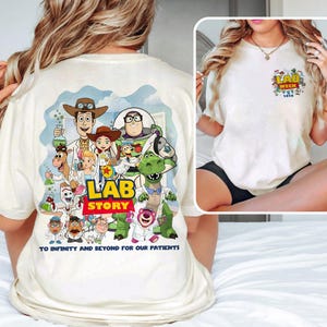 May include: Cream-colored t-shirt featuring a colorful graphic of Toy Story characters in lab coats, with the text "LAB STORY" and "TO INFINITY AND BEYOND FOR OUR PATIENTS." A smaller graphic on the front pocket reads "LAB WEEK 2016."