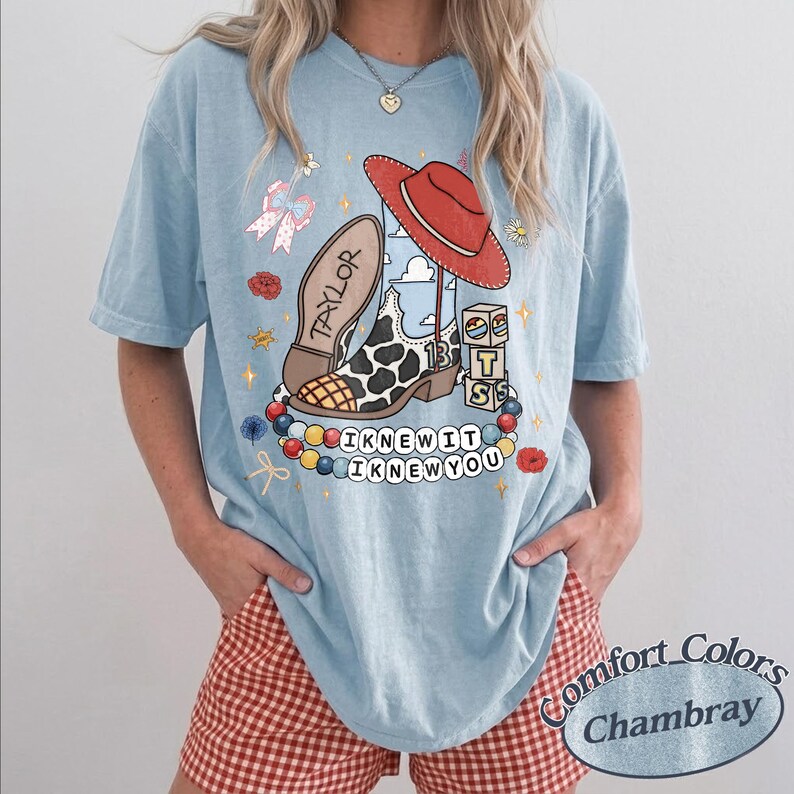 May include: A light blue t-shirt featuring a graphic print of a cow-print cowboy boot with the name TAYLOR, a red cowboy hat, alphabet blocks, and a string of colourful beads. The text I KNEW IT I KNEW YOU is printed below the graphic design.