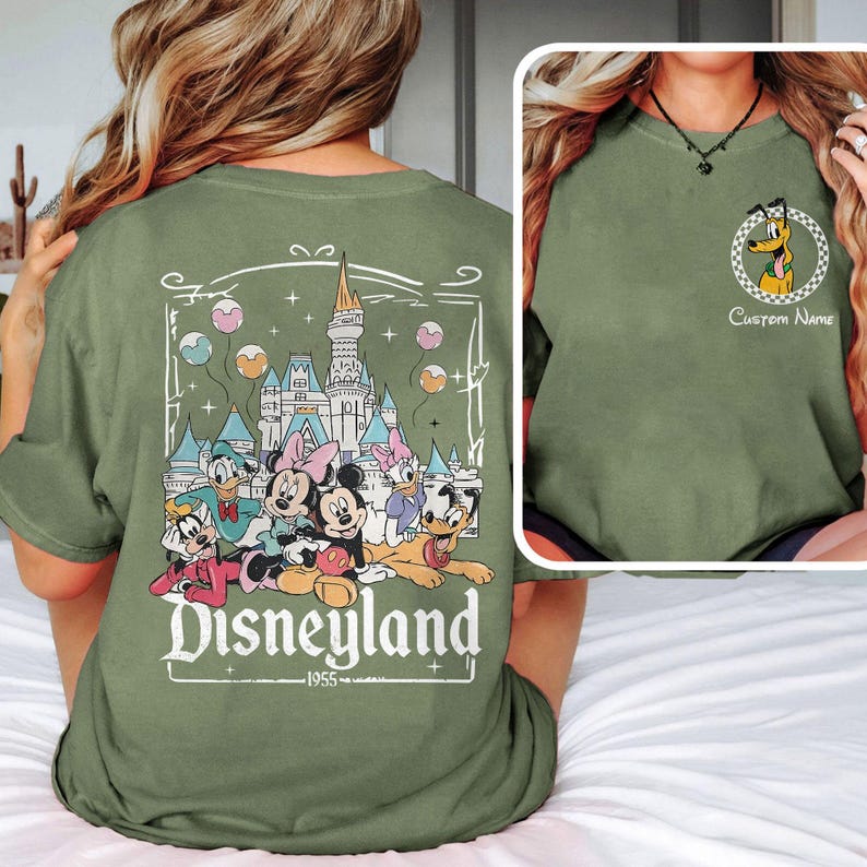 May include: Green t-shirt with a graphic of Mickey Mouse and friends in front of the Sleeping Beauty Castle at Disneyland. The text "Disneyland 1955" is below the graphic.