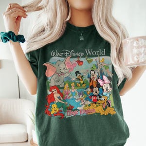 Pode incluir: T-shirt verde escuro com um gr&aacute;fico colorido de v&aacute;rios personagens da Disney, incluindo Mickey Mouse, Minnie Mouse e Dumbo. As palavras "Walt Disney World" e "Magic Kingdom" s&atilde;o impressas na camisa.