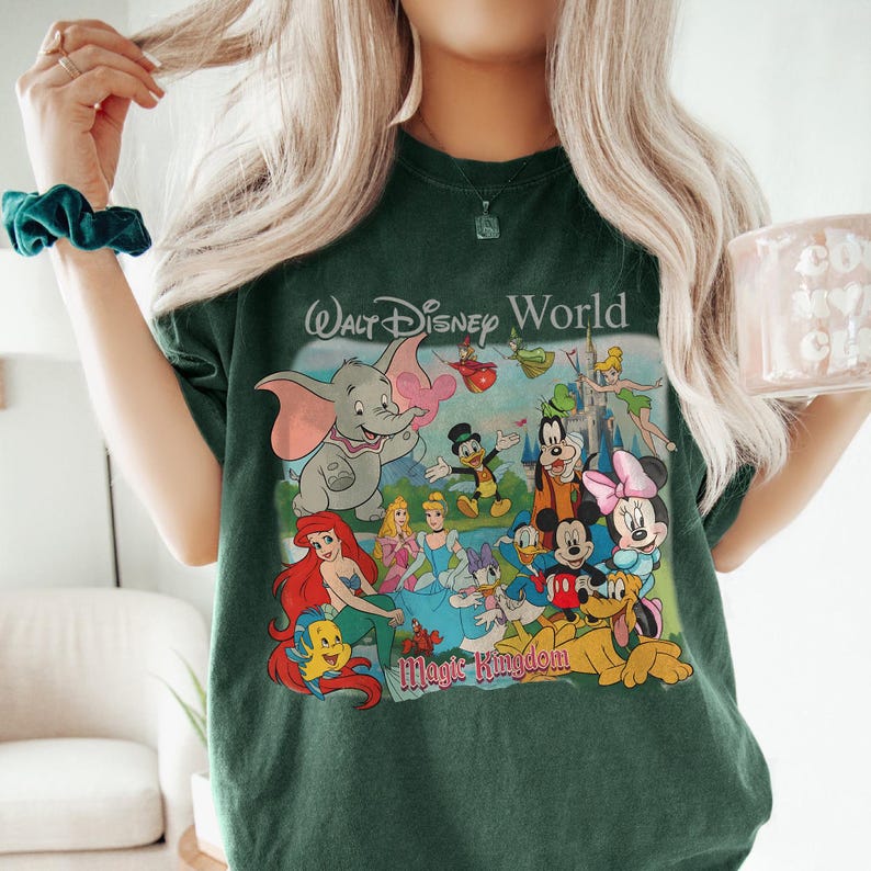 Pode incluir: T-shirt verde escuro com um gr&aacute;fico colorido de v&aacute;rios personagens da Disney, incluindo Mickey Mouse, Minnie Mouse e Dumbo. As palavras "Walt Disney World" e "Magic Kingdom" s&atilde;o impressas na camisa.