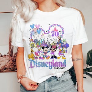May include: White t-shirt featuring a colorful graphic celebrating Disneyland's 70th anniversary. The design includes Mickey and Minnie Mouse, Donald and Daisy Duck, Goofy, Pluto, and a castle, with the text "Disneyland 1955-2025".