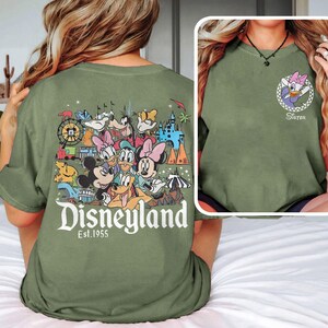 May include: Olive green t-shirt with a Disneyland graphic on the back featuring Mickey Mouse, Minnie Mouse, Donald Duck, and other Disney characters. The front has a Daisy Duck graphic and the word "Sister".