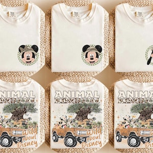 May include: Cream-colored t-shirts featuring Mickey Mouse and Goofy in safari hats, and the text "Animal Kingdom Wild about Disney." The shirts are folded on woven mats, showcasing a theme of adventure and Disney characters.