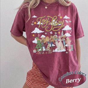 May include: A berry-coloured short-sleeve t-shirt featuring a graphic print of Toy Story characters, white clouds, stars, and musical notes. The text Toy Story is written in a yellow rope-style font across the top. The shirt has a crew neckline.