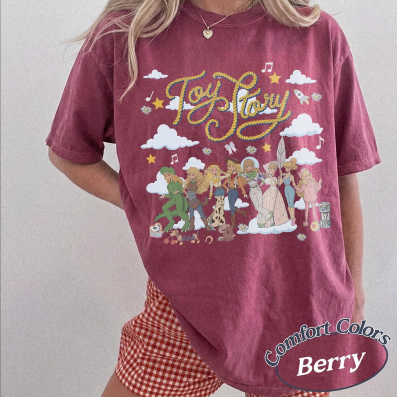 May include: A berry-coloured short-sleeve t-shirt featuring a graphic print of Toy Story characters, white clouds, stars, and musical notes. The text Toy Story is written in a yellow rope-style font across the top. The shirt has a crew neckline.