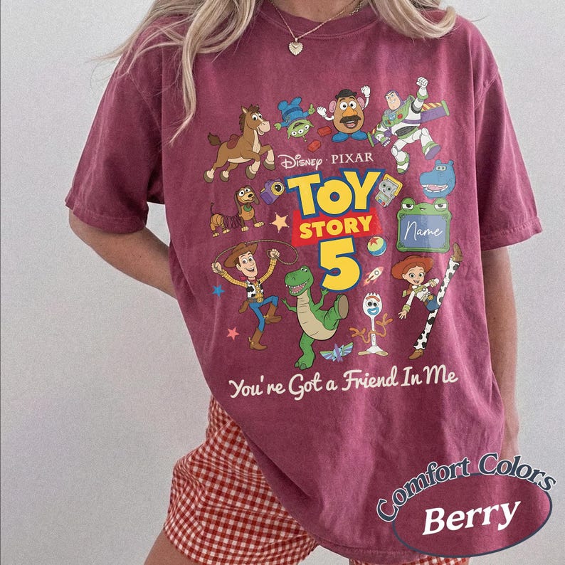 May include: A berry-colored t-shirt featuring a graphic print of Disney Pixar Toy Story characters including Woody, Buzz Lightyear, Rex, and Mr. Potato Head. The text reads Toy Story 5 and You've Got a Friend In Me, with a space for a name.