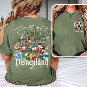 May include: Olive green t-shirt featuring a Disneyland graphic with Mickey Mouse, Donald Duck, Goofy, and Pluto in Santa hats. The back of the shirt displays a Christmas scene with a castle and the text "Disneyland 1955".
