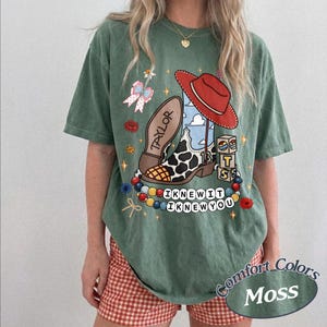 May include: A moss green short-sleeve t-shirt featuring a graphic print of a cow-print cowboy boot with a red hat, blocks labelled "TS", and a string of beads with the text "I KNEW IT I KNEW YOU". The boot sole is inscribed with "TAYLOR".