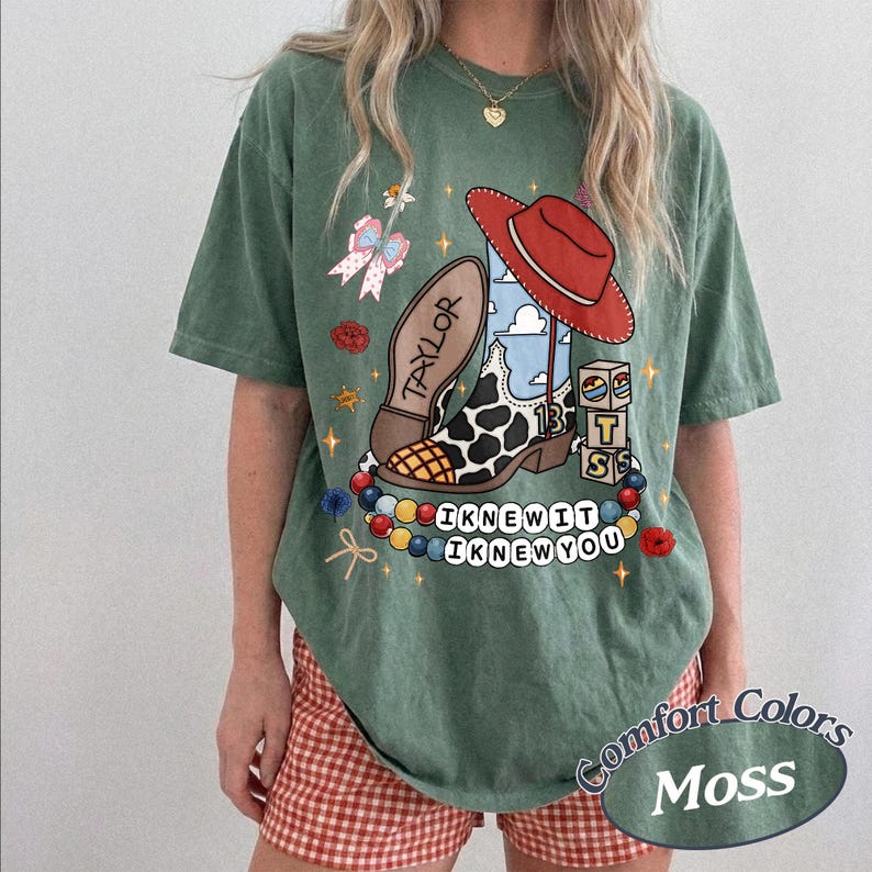 May include: A moss green short-sleeve t-shirt featuring a graphic print of a cow-print cowboy boot with a red hat, blocks labelled "TS", and a string of beads with the text "I KNEW IT I KNEW YOU". The boot sole is inscribed with "TAYLOR".