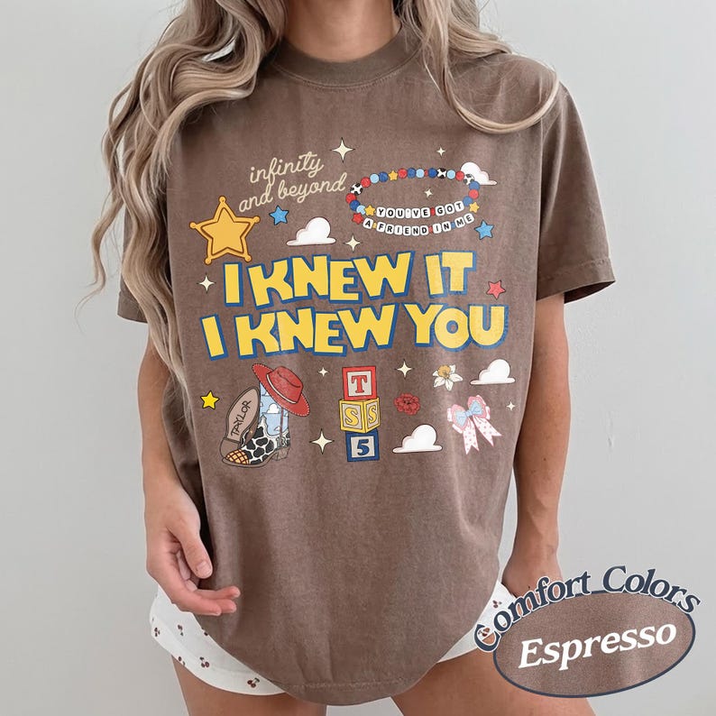May include: A brown short-sleeve t-shirt featuring a graphic print with the text "infinity and beyond", "YOU'VE GOT A FRIEND IN ME", "I KNEW IT", and "I KNEW YOU". The design includes a sheriff star, a beaded bracelet, a cowboy boot, and blocks.