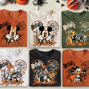 Op de afbeelding: Zes verschillende T-shirts met een Halloween-thema met Mickey Mouse, Minnie Mouse, Goofy en Donald Duck. De shirts zijn oranje, wit, zwart en groen. De shirts hebben de tekst "Boo!" en "Halloween" erop.