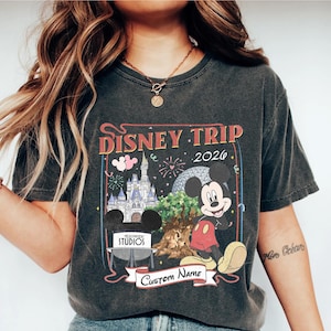 Pu&ograve; includere: T-shirt grigio scuro con una grafica colorata di Disney Trip 2026 con Topolino, un castello e fuochi d'artificio. Il design include il testo "Hollywood Studios" e uno striscione per un nome personalizzato.