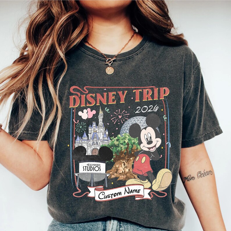 Pu&ograve; includere: T-shirt grigio scuro con una grafica colorata di Disney Trip 2026 con Topolino, un castello e fuochi d'artificio. Il design include il testo "Hollywood Studios" e uno striscione per un nome personalizzato.