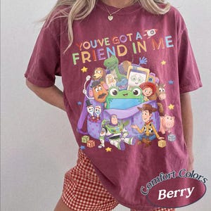 May include: A berry-colored t-shirt featuring a graphic print of various animated characters from the Toy Story franchise emerging from a purple backpack, with the text "YOU'VE GOT A FRIEND IN ME" printed in colorful letters above the characters.