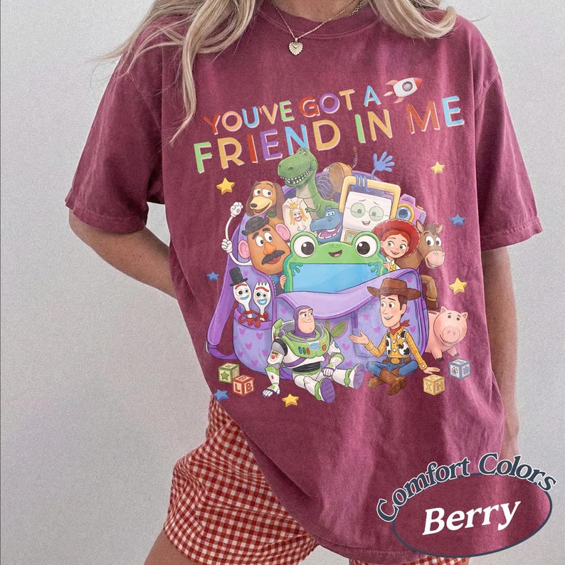 May include: A berry-colored t-shirt featuring a graphic print of various animated characters from the Toy Story franchise emerging from a purple backpack, with the text "YOU'VE GOT A FRIEND IN ME" printed in colorful letters above the characters.