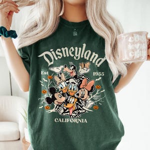 K&ouml;nnte beinhalten: Gr&uuml;nes T-Shirt mit einem Vintage-Disneyland-Motiv, das Mickey Mouse, Minnie Mouse, Donald Duck, Goofy und Pluto in Skelettkost&uuml;men zeigt. Der Text lautet "Disneyland Est 1955 California".