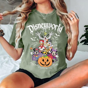 May include: An olive green t-shirt with a Halloween-themed Disney World graphic. The design features various Disney characters dressed in costumes, including Mickey Mouse, Goofy, and Donald Duck, with a large jack-o'-lantern in the center. The text "Disneyworld" is at the top.