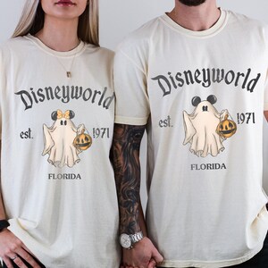 May include: Ivory-colored t-shirts with a Disneyworld ghost design. The design includes the text "Disneyworld est. 1971 FLORIDA" and a cartoon ghost holding a jack-o'-lantern. The ghost has Mickey Mouse ears. The shirts are short-sleeved.