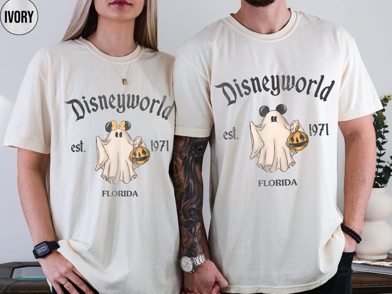 May include: Ivory-colored t-shirts with a Disneyworld ghost design. The design includes the text "Disneyworld est. 1971 FLORIDA" and a cartoon ghost holding a jack-o'-lantern. The ghost has Mickey Mouse ears. The shirts are short-sleeved.