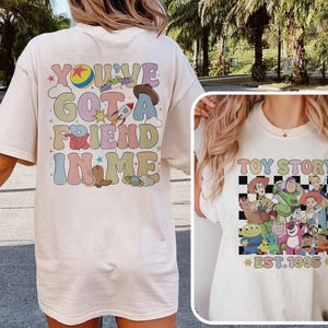 May include: White t-shirt with a colorful graphic on the back that says "You've Got a Friend in Me" with a cartoon rocket, cowboy hat, and stars. The front of the shirt has a black and white checkered pattern with cartoon characters from Toy Story and the text "Toy Story Est. 1995".