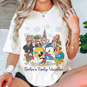 Może przedstawiać: Kremowa koszulka z kolorową grafiką postaci Disneya, numerem "2026" i napisem "Smith's Family Vacation". Projekt zawiera Myszka Miki, Kaczor Donald i Goofy.