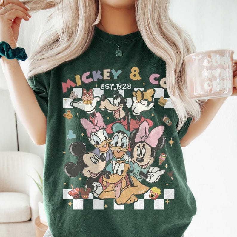 May include: Dark green t-shirt featuring a graphic of Mickey Mouse, Minnie Mouse, Donald Duck, Daisy Duck, and Pluto. The shirt has the text "Mickey & Co. Est. 1928" in a retro style, with a checkered background.