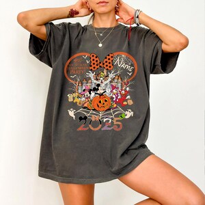 May include: Dark grey t-shirt with a Halloween-themed design. It features Mickey and Minnie Mouse, a pumpkin, and the text "Halloween Party 2025." The design includes a castle, ghosts, and a spider web.