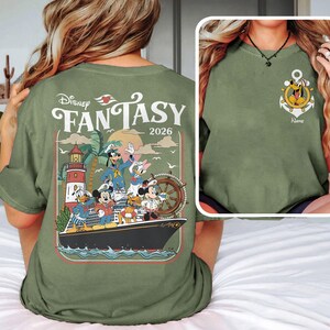 Puede incluir: Camiseta verde oliva con un dise&ntilde;o de Disney Fantasy 2026 en la espalda. El gr&aacute;fico incluye personajes de Disney en un barco, un faro y palmeras. La parte delantera tiene un dise&ntilde;o de ancla con Pluto.