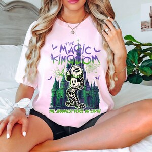 May include: Pale pink t-shirt with a Halloween design. The graphic shows a skeleton Mickey Mouse holding a skull, a castle, bats, and the text "The Magic Kingdom" and "The Spookiest Place on Earth" in green and purple.