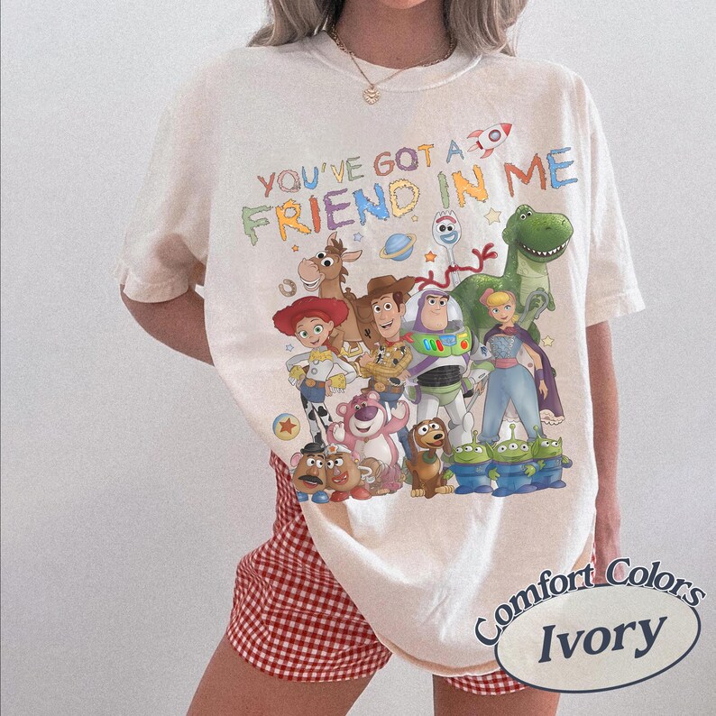 May include: An ivory short-sleeve t-shirt featuring a colorful graphic print of various animated characters from the Toy Story franchise. The text "YOU'VE GOT A FRIEND IN ME" is printed in a multi-colored font above the group of characters.