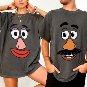 May include: Two charcoal grey t-shirts featuring cartoon faces. One shirt has a female face with pink lips and nose, and the other has a male face with a moustache and orange nose. The shirts are a matching set.