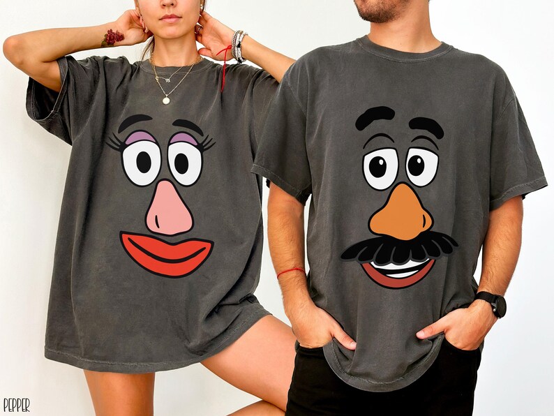 May include: Two charcoal grey t-shirts featuring cartoon faces. One shirt has a female face with pink lips and nose, and the other has a male face with a moustache and orange nose. The shirts are a matching set.