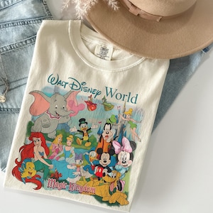 Pode incluir: Camiseta creme com um gr&aacute;fico colorido de personagens da Disney, incluindo Dumbo, Mickey Mouse e Ariel. A camisa tem o texto "Walt Disney World" e "Magic Kingdom" em fonte azul. Um chap&eacute;u bege e jeans azuis est&atilde;o no fundo.
