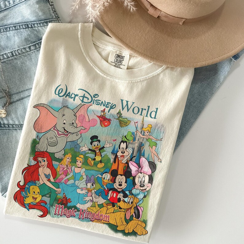 Pode incluir: Camiseta creme com um gr&aacute;fico colorido de personagens da Disney, incluindo Dumbo, Mickey Mouse e Ariel. A camisa tem o texto "Walt Disney World" e "Magic Kingdom" em fonte azul. Um chap&eacute;u bege e jeans azuis est&atilde;o no fundo.