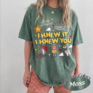 May include: A moss green short-sleeve t-shirt featuring a graphic print with the text "infinity and beyond", "I KNEW IT I KNEW YOU", and "YOU'VE GOT A FRIEND IN ME". The design includes a yellow star, a beaded bracelet, a cowboy boot, and blocks.