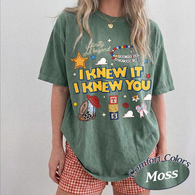 May include: A moss green short-sleeve t-shirt featuring a graphic print with the text "infinity and beyond", "I KNEW IT I KNEW YOU", and "YOU'VE GOT A FRIEND IN ME". The design includes a yellow star, a beaded bracelet, a cowboy boot, and blocks.