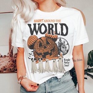 May include: White t-shirt with a vintage-style graphic featuring a pumpkin-shaped Mickey Mouse head, a castle, and ghosts. The text "Haunt Around the World" is printed in black above the graphic. The text "It's really Not-So-Scary!" is printed in black below the graphic.