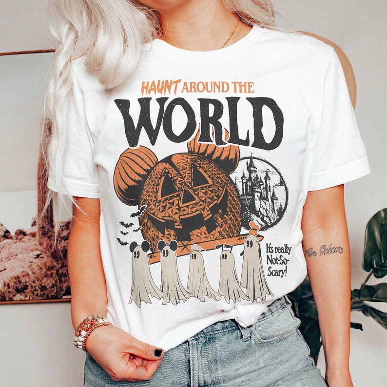 May include: White t-shirt with a vintage-style graphic featuring a pumpkin-shaped Mickey Mouse head, a castle, and ghosts. The text "Haunt Around the World" is printed in black above the graphic. The text "It's really Not-So-Scary!" is printed in black below the graphic.