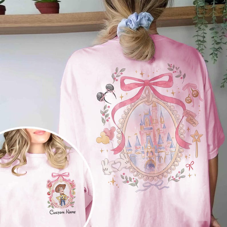 Pode incluir: Camiseta rosa claro com um design fantasioso com um castelo, orelhas do Mickey Mouse e um la&ccedil;o rosa. A camiseta tamb&eacute;m tem uma imagem menor de um personagem de desenho animado com o texto "Custom Name".