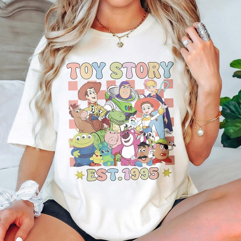 May include: White t-shirt with a colorful graphic of characters from the movie Toy Story. The graphic includes Woody, Buzz Lightyear, Jessie, and other characters. The text "TOY STORY EST. 1995" is printed below the graphic.