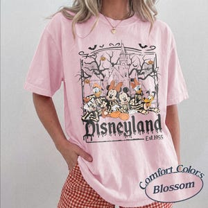 May include: Pink t-shirt with a Disneyland Halloween graphic. The design features Mickey and Minnie Mouse, Donald Duck, and Goofy in skeleton costumes in front of a haunted castle. The text "Disneyland Est. 1955" is below the graphic.