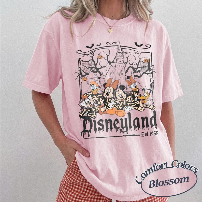 May include: Pink t-shirt with a Disneyland Halloween graphic. The design features Mickey and Minnie Mouse, Donald Duck, and Goofy in skeleton costumes in front of a haunted castle. The text "Disneyland Est. 1955" is below the graphic.