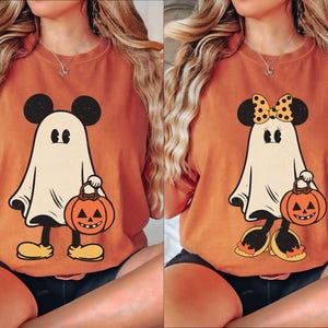 May include: Two orange Halloween themed t-shirts with a ghost Mickey Mouse design. One shirt has a black and white polka dot bow on the ghost's head. Both ghosts are holding a jack-o-lantern.