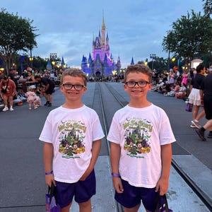 May include: Two young people wearing white t-shirts with a Halloween design, purple shorts, and glasses stand in front of a castle. The shirts read "Mickey's Not-So-Scary Halloween Party" with a graphic of Mickey Mouse and friends. They each hold a purple bag.