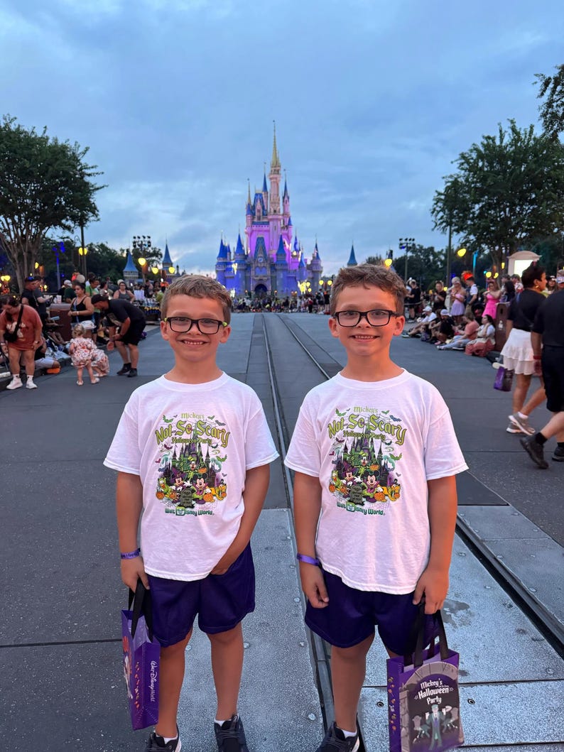 May include: Two young people wearing white t-shirts with a Halloween design, purple shorts, and glasses stand in front of a castle. The shirts read "Mickey's Not-So-Scary Halloween Party" with a graphic of Mickey Mouse and friends. They each hold a purple bag.