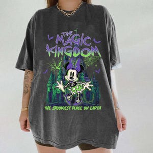 May include: Dark gray t-shirt featuring a spooky Minnie Mouse design in green, purple, and white. The text "THE MAGIC KINGDOM" and "THE SPOOKIEST PLACE ON EARTH" are printed above and below the graphic. The shirt has short sleeves.