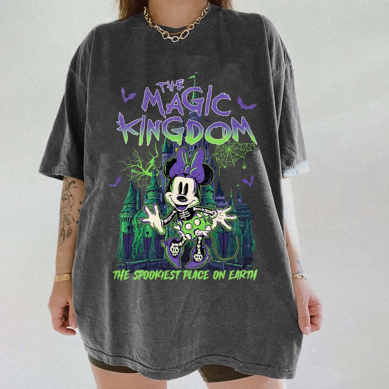 May include: Dark gray t-shirt featuring a spooky Minnie Mouse design in green, purple, and white. The text "THE MAGIC KINGDOM" and "THE SPOOKIEST PLACE ON EARTH" are printed above and below the graphic. The shirt has short sleeves.