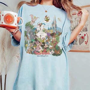 K&ouml;nnte beinhalten: Hellblaues T-Shirt mit einem farbenfrohen Aufdruck von Disney-Figuren, darunter Mickey Mouse und Dumbo. Der Text "Magic Kingdom Walt Disney World" ist darunter gedruckt. Eine wei&szlig;e und orangefarbene Tasse mit einem Sonnendesign wird im Bild gehalten.